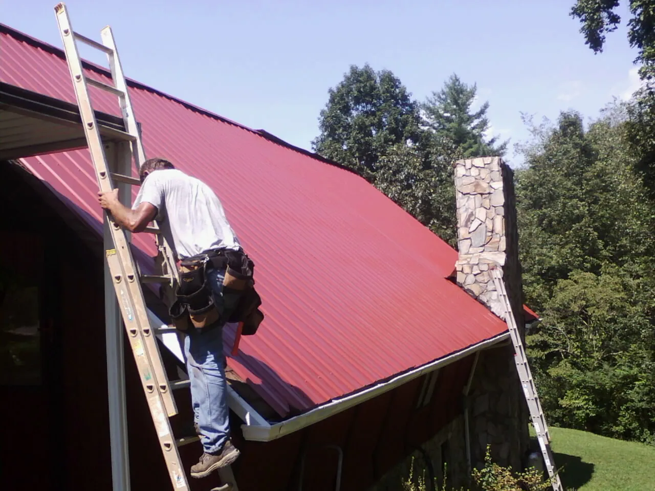 Roofer on a ladder beside a red metal roof and stone chimney — Soffit & Fascia Repair by Roofing Brothers in Archer